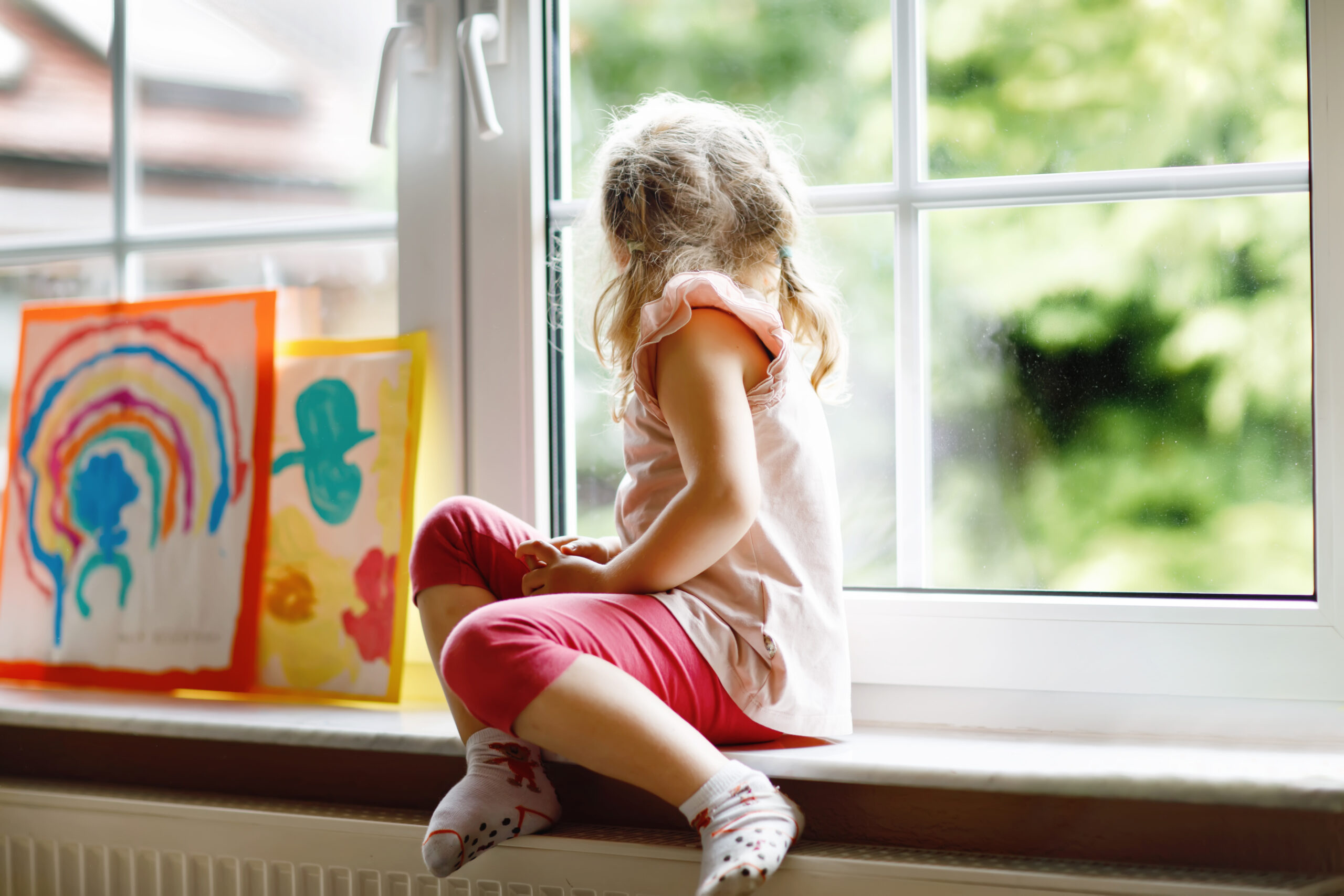 White toddler girl by window with rainbow painted with colorful colors. 